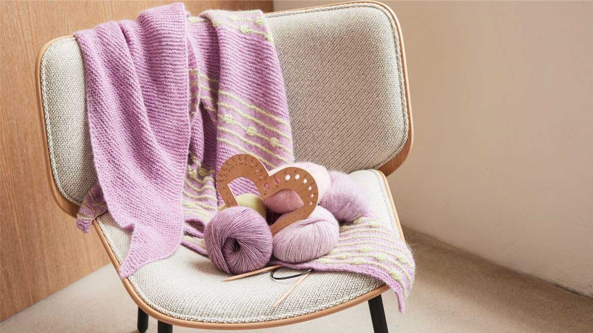 gestrickte Tücher rosafarben mit weissem Muste , davor Wollknaeuel rosa mit Nadelmesser aus Holz
